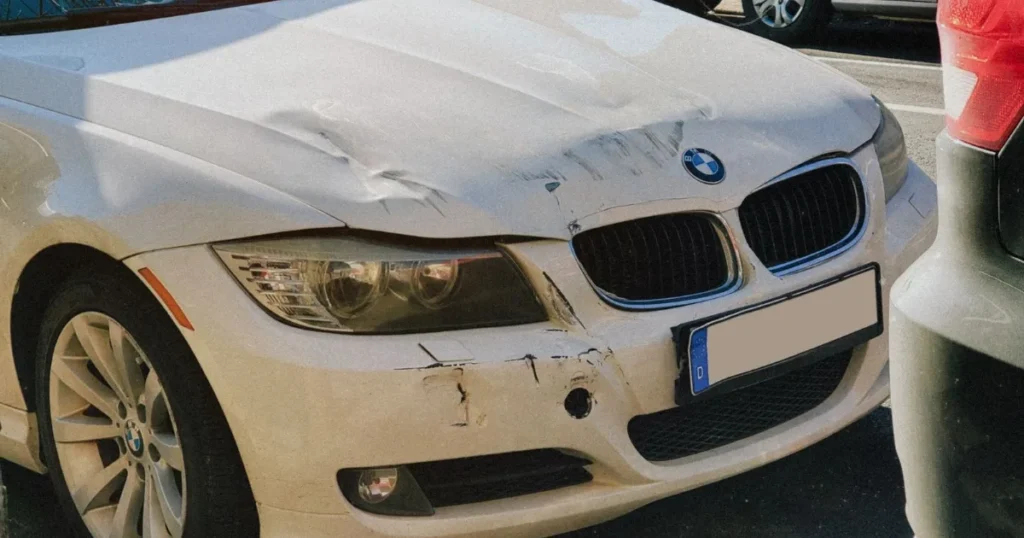 White sedan with crumpled front-end damage parked at the side of a city street after a collision