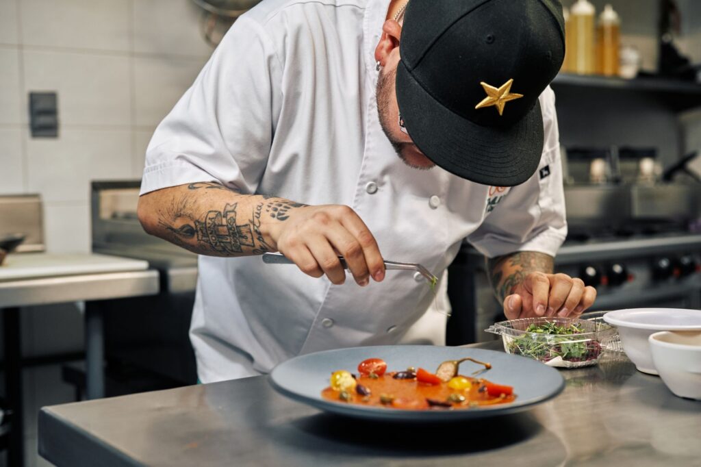 A cook plating a dish on a stainless steel pass in a commercial restaurant kitchen
